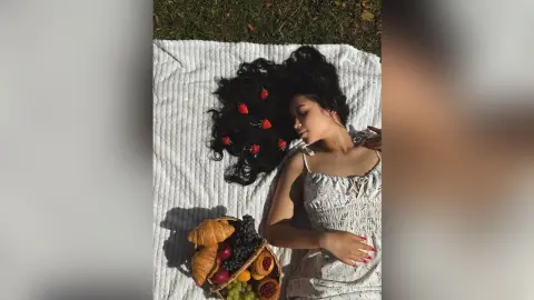 MinnieMile lies on a white blanket with her long black hair spread out, surrounded by strawberries and a basket of fruit.