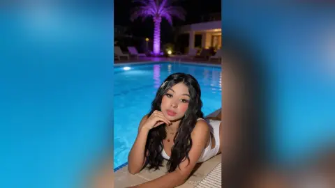 Young woman in white swimwear relaxing by a glowing blue swimming pool at a tropical night resort.