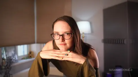 Young woman with glasses in an olive green top looking calm in a cozy, sunlit indoor setting.