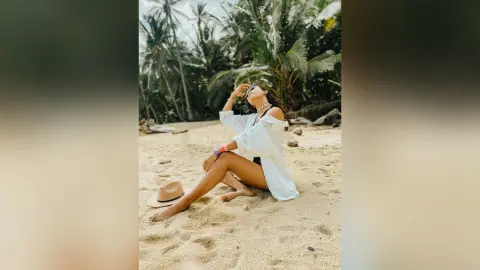 Woman in white linen shirt sitting on a tropical beach under palm trees during a relaxing summer vacation.