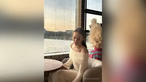 GwendolynBucey wearing a white off-the-shoulder long-sleeve top, sitting by a window overlooking a body of water at sunset.