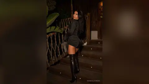 BiankaLeoness poses in a tight black mini dress and tall black boots while standing on outdoor stairs at night.