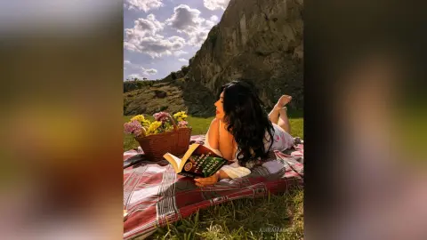 Relaxed woman in white loungewear reading a book on a picnic blanket near a mountain under a sunny sky.