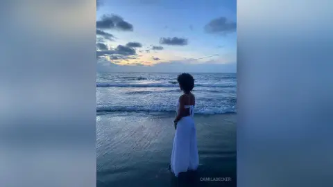 Woman in a white summer dress standing on a beach at twilight looking at the ocean waves.
