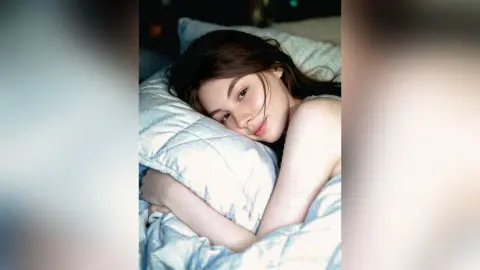 Young woman with long brown hair relaxing on white pillows in a cozy, peaceful bedroom setting.