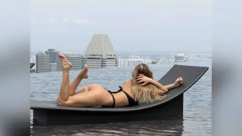Relaxed blonde woman in black bikini lounging on a rooftop sunbed overlooking a city skyline at dusk.