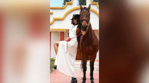 Elegant woman in a white maxi dress and black hat standing next to a brown horse in a sunny courtyard.