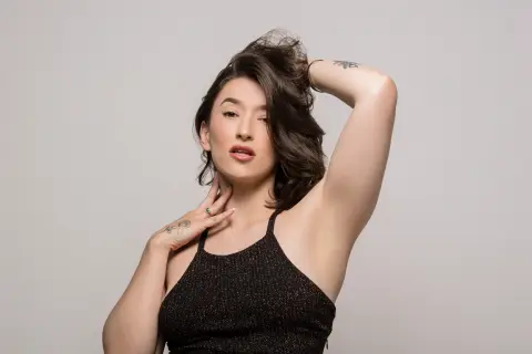 Katt_Henao poses with one hand in her dark hair and the other near her neck, wearing a black halter top against a plain grey background.
