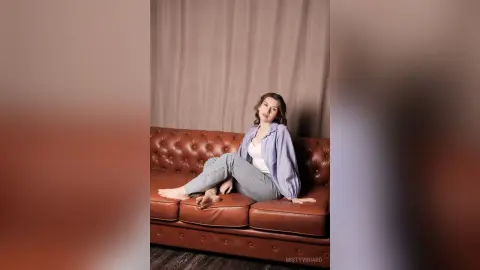 Young woman in a light blue shirt and jeans relaxing on a brown leather sofa in a minimalist studio setting.