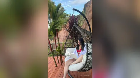 Relaxed woman in a white cover-up reading a book in a hanging egg chair on a tropical wooden deck.