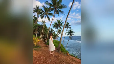 Dreamy portrait of a woman in a white summer dress standing by palm trees on a tropical beach at sunset.