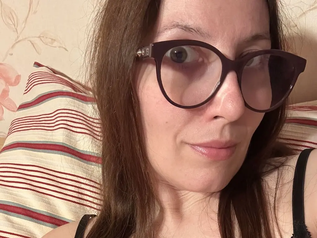 Smiling woman with long brown hair wearing stylish black glasses relaxing on a striped pillow indoors.
