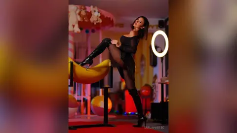 Glamorous woman in a black mini dress and tall boots posing in a colorful, playful room with soft lighting.