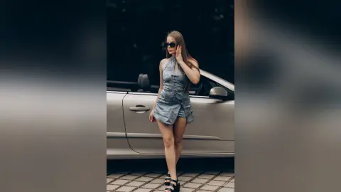 AdelinaKael poses in a denim halter-neck romper and sunglasses, standing next to a white car outdoors.