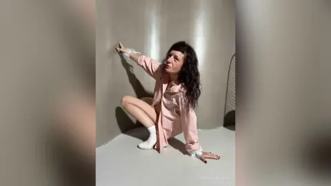 SabrinaBauer — SabrinaBauer poses in a pink button-down shirt and white socks against a grey wall.