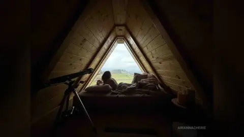 Woman relaxing in a cozy wooden A-frame cabin bedroom with a view of green hills through a triangular window.