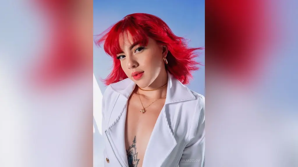 AnaKozlov β Young woman with vibrant red hair wearing a white blazer and gold necklace against a blue sky background.