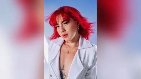 Young woman with vibrant red hair wearing a white blazer and gold necklace against a blue sky background.
