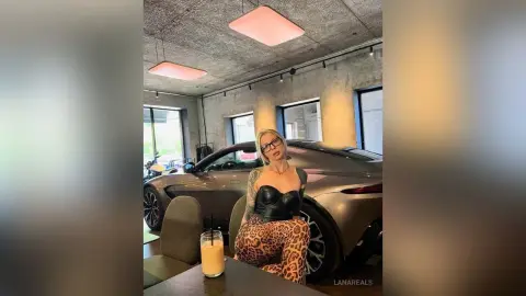 Stylish woman in a black corset and leopard print leggings posing confidently next to a luxury car indoors.