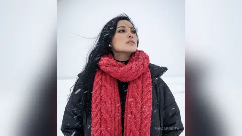 Serene woman with long dark hair wearing a cozy red knit scarf and black winter coat in a snowy landscape.