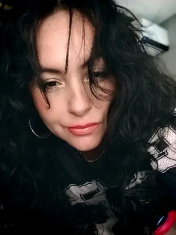 Smiling woman with wavy dark hair wearing a black and white patterned top in a soft indoor setting.