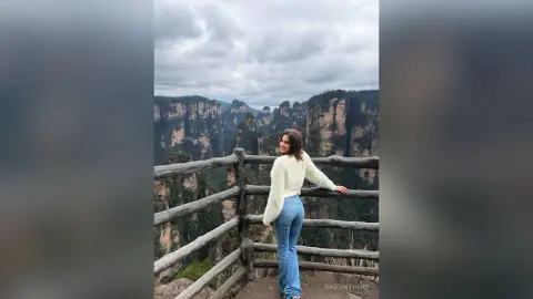 Woman in a white fuzzy sweater and blue jeans standing on a wooden balcony overlooking misty mountain cliffs.