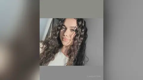 Young woman with long wavy brunette hair wearing a white ribbed top in soft natural sunlight.