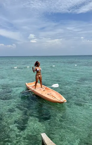 Woman in white bikini standing on an orange kayak in turquoise tropical ocean water under a blue sky.