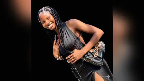 Smiling woman with long braids wearing a black sleeveless dress and designer crossbody bag outdoors.