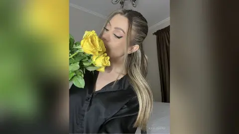 Blonde woman in a black satin shirt smelling a bouquet of yellow roses in a cozy indoor setting.