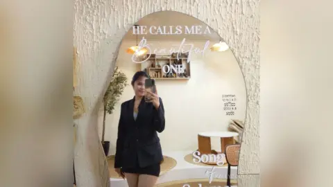 Woman in a black blazer taking a mirror selfie in a bright, modern minimalist room.
