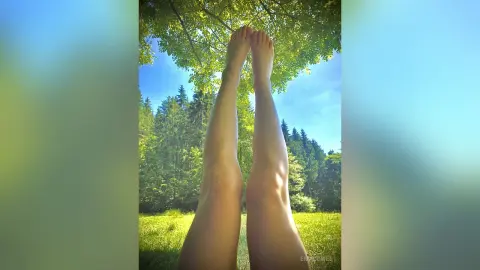 EmaLowee shows her long, bare legs stretched up toward the green leaves of a tree in a sunny forest setting.
