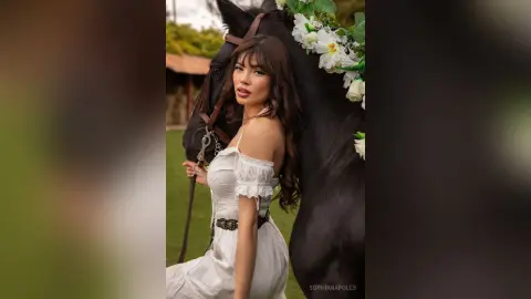 Elegant woman in a white summer dress posing with a black horse in a lush green garden setting.