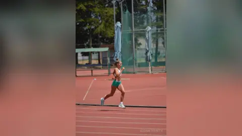 Athletic woman in green sports bra and shorts running on a red track field during a sunny outdoor workout.