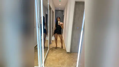 Stylish woman in a black mini dress taking a mirror selfie in a modern, brightly lit hallway.