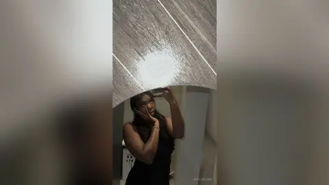 AmyJacobs poses in a black top, touching her face while looking into a mirror in a bathroom setting.