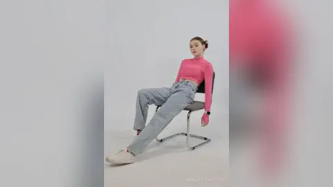 Young woman in pink turtleneck and light blue jeans sitting on a chair against a white studio background.