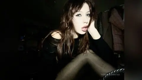 Moody portrait of a brunette woman in a black long-sleeve top posing in a dimly lit room.