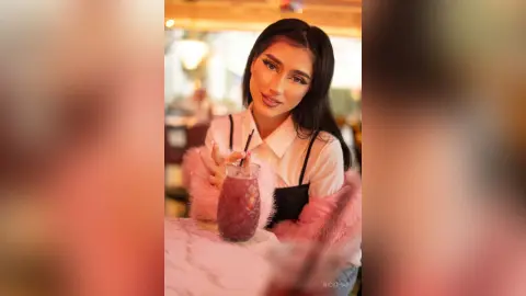 Young woman in a white blouse and pink fuzzy cardigan enjoying a berry smoothie in a warm cafe setting.