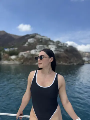 Woman in black one-piece swimsuit and sunglasses relaxing on a boat in the ocean under a blue sky.