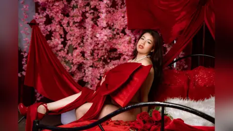 Elegant woman in a red gown reclining on a bed amidst pink cherry blossoms and romantic red drapery.