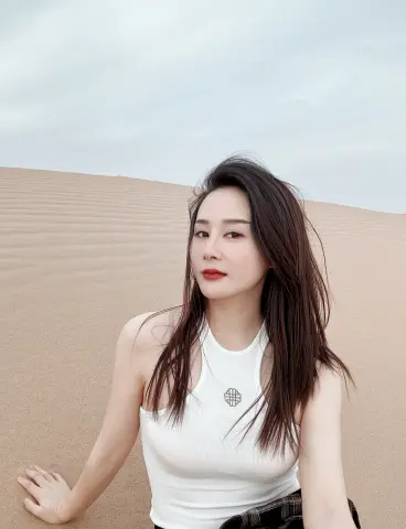 Young woman in a white ribbed tank top sitting on desert sand dunes under a cloudy sky.