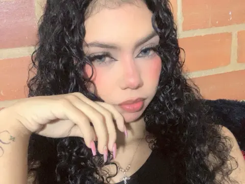 Nathy_Pelaez poses with her hand under her chin, showing her long curly black hair and light pink blush against a brick wall background.