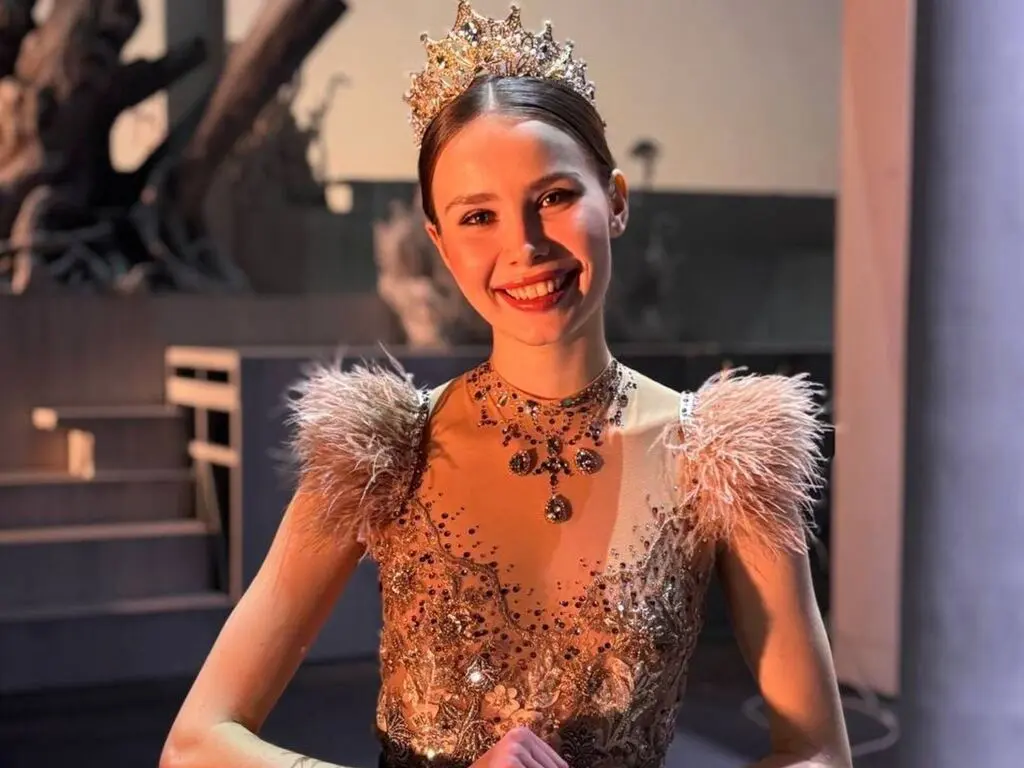 Smiling young woman in a glamorous gold sequin gown and crown posing in a dramatic indoor setting.