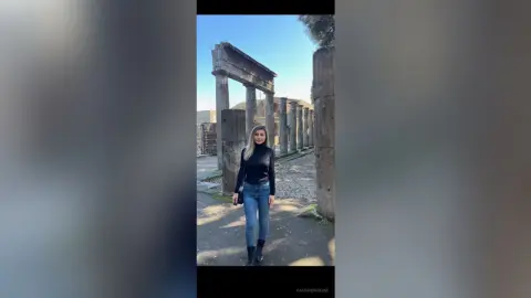 Woman in black turtleneck and jeans standing amidst ancient stone ruins under a clear blue sky.