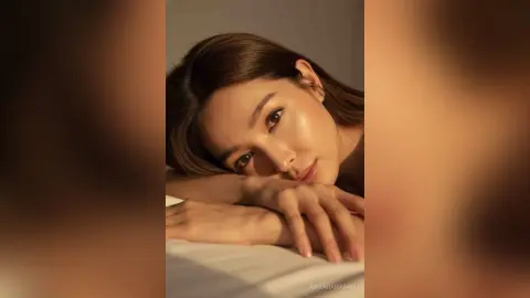 Close-up portrait of a woman with glowing skin resting her head on her arms in warm, soft lighting.