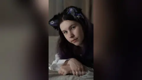 Young woman with purple hair wearing black cat ears in a moody indoor setting.