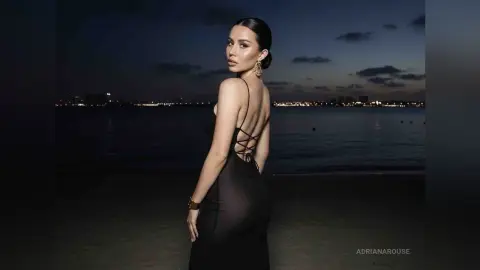 Elegant woman in a black backless evening gown posing by the ocean at night under city lights.