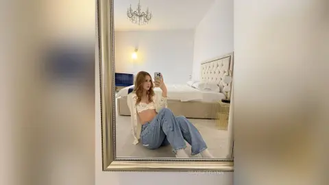 Young woman in a white lace bralette and baggy jeans taking a mirror selfie in a bright, modern bedroom.