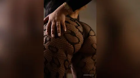 Curvy woman in black lace patterned tights posing in a moody, dimly lit indoor setting.